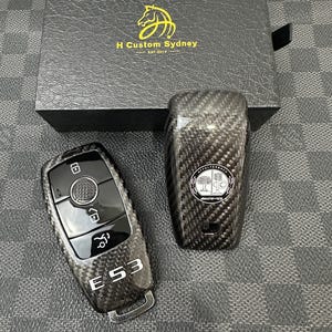 May include: Two carbon fiber car key fobs with a black box in the background. One fob has the text "E 53" and the other has the AMG logo. The box has the text "H Custom Sydney EST. 2019" and a horse logo.