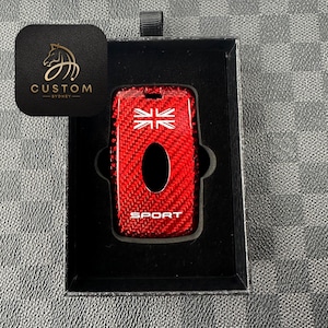 Puede incluir: Una funda roja de fibra de carbono para la llave del coche con un diseño de la Union Jack y la palabra "SPORT". La llave está en una caja negra con una etiqueta negra con el logotipo "CUSTOM SYDNEY".
