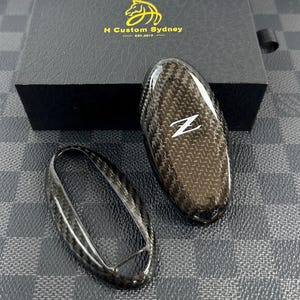May include: Two black carbon fiber key covers for a car key. The covers are shaped like a key fob and have a carbon fiber pattern. One cover has the letter "Z" printed on it.