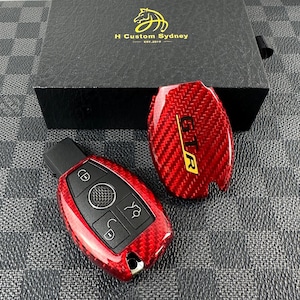May include: Red carbon fiber key fob cover with a black key fob and four buttons. The key fob cover has the letters "GT" in gold lettering. The key fob is on a black and white checkered surface.