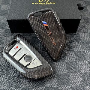 Puede incluir: Dos cubiertas de llave de coche de fibra de carbono negra. Una cubierta tiene un botón plateado con un icono rojo y la otra cubierta tiene el texto "M3 CS" en letras plateadas.