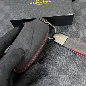 May include: Black key fob cover with red stitching and a matching key chain. The key fob is made of a suede-like material and has a metal key ring. The key chain has a leather strap with red stitching and a metal clasp.