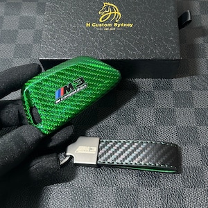 Könnte beinhalten: Eine grüne Autoschlüsselhülle aus Carbon mit M3 Competition Logo und ein passender schwarzer Carbon-Schlüsselanhänger mit M3-Metallemblem, präsentiert in einer schwarzen, strukturierten Box mit der Aufschrift H Custom Sydney Est. 2019.