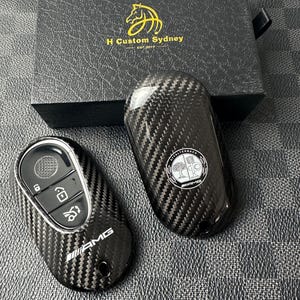 Puede incluir: Dos llaveros de coche de fibra de carbono con un patrón de tejido texturizado. Un llavero tiene el logotipo de AMG y el otro el logotipo de Mercedes-Benz. La caja detrás de los llaveros tiene el texto "H Custom Sydney EST. 2019".