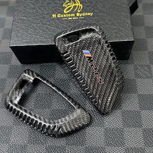 Puede incluir: Funda de fibra de carbono para la llave del coche con el texto "M2 CS" y el logotipo de BMW. La funda de la llave es negra y tiene una superficie texturizada. La funda de la llave está junto a otra funda de llave a juego y una caja negra con texto dorado.