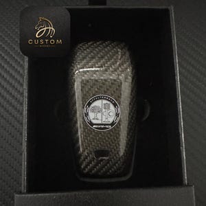 Puede incluir: Un llavero de fibra de carbono negro con un logotipo Mercedes-Benz AMG plateado y el texto "Affalterbach" alrededor del logotipo.