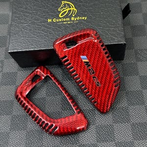 Puede incluir: Funda roja de fibra de carbono para llavero con el texto "M2 CS" y una franja multicolor. La funda del llavero está junto a un marco de llavero a juego. La caja del fondo tiene el texto "H Custom Sydney".