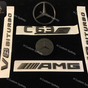 Puede incluir: Emblemas negros de Mercedes-Benz, incluyendo un logotipo de estrella de tres puntas, "C63S", "V8 BITURBO" y la inscripción "AMG". Los emblemas están sobre un fondo blanco.
