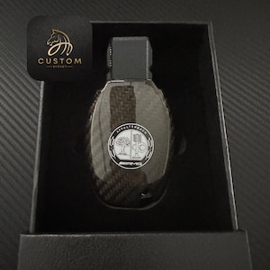 May include: A black carbon fiber key fob with a silver AMG logo and the text "Affalterbach" around the logo. The key fob is in a black box.