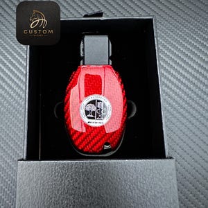 May include: A red car key fob with a carbon fibre pattern and a silver Mercedes-Benz AMG logo. The key fob is in a black box.