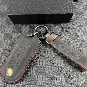 Puede incluir: Un llavero de cuero negro con detalles de costura roja. El llavero tiene un anillo de metal y un llavero de cuero negro con la palabra "GTS" en relieve.
