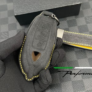 Könnte beinhalten: Ein schwarzer Autoschlüsselanhänger mit passendem Schlüsselanhänger. Der Schlüsselanhänger hat drei Knöpfe und ein Logo. Der Schlüsselanhänger und der Schlüsselanhänger sind mit gelben Nähten versehen. Das Wort "Performante" ist sichtbar.