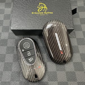 May include: Two carbon fiber car key fobs with a black box in the background. The key fobs have buttons for locking, unlocking, and other functions. The box has a gold horse logo and the text "H Custom Sydney EST. 2019".