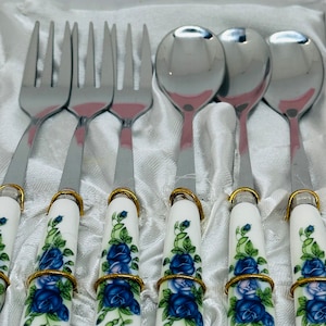 May include: Set of six silver-toned teaspoons and forks with white handles decorated with blue roses and gold accents. The handles are in a velvet-lined box.