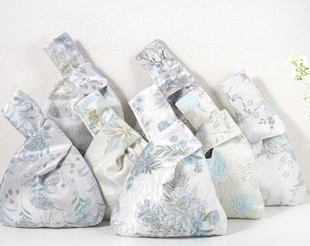 May include: A collection of five Japanese-style knot bags made from fabric with various floral and botanical patterns in shades of light blue, white, and silver. Each bag features a unique asymmetrical handle design with one strap longer than the other.