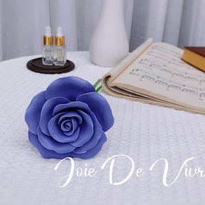 Puede incluir: Una sola rosa de tela azul grande con un tallo verde. La rosa está sobre una superficie blanca con el texto "Joie De Vivre" escrito debajo.