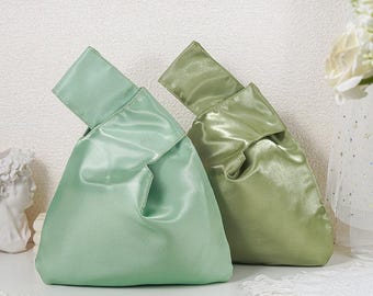May include: Two wristlet bags made of a smooth, shiny satin fabric. One bag is a light mint green color, and the other is a slightly darker olive green. Both bags feature a single integrated handle loop and a soft, unstructured silhouette.