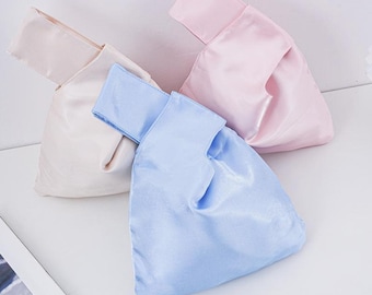 May include: Three small wristlet bags made of a smooth, satin-like fabric. One bag is light blue, one is pale pink, and one is a soft cream color. Each bag features a unique Japanese-style knot handle design with one strap longer than the other.