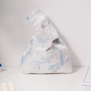 May include: A white knot bag featuring a crane design in blue and pink. The bag has a pink interior and a decorative knot handle. The bag is made of a silky material, ideal for carrying small items.