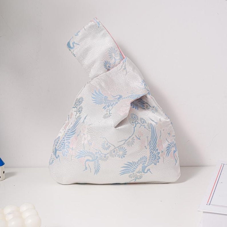 May include: A white knot bag featuring a crane design in blue and pink. The bag has a pink interior and a decorative knot handle. The bag is made of a silky material, ideal for carrying small items.
