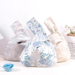 May include: Three fabric knot bags in the image. The bags have a rounded shape with a handle. One bag is light gray with a floral pattern, another is white with blue flowers, and the third is cream-colored with a lace design.