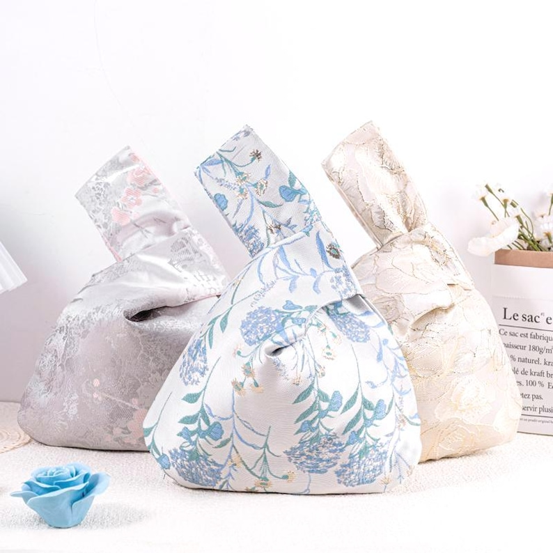 May include: Three fabric knot bags in the image. The bags have a rounded shape with a handle. One bag is light gray with a floral pattern, another is white with blue flowers, and the third is cream-colored with a lace design.