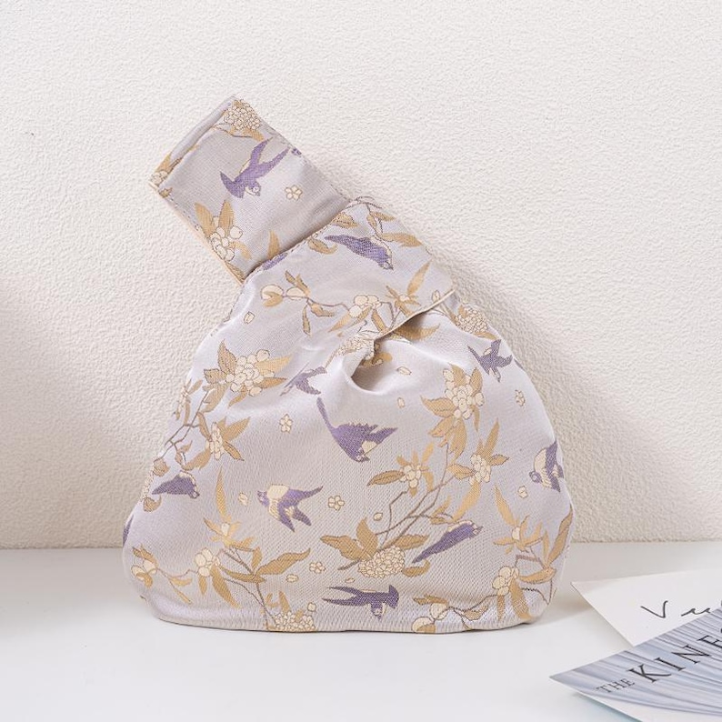 May include: A light grey knotted handbag with a floral and bird pattern. The design features gold and purple birds and flowers on a light grey background. The bag is made of a soft, possibly satin-like material.