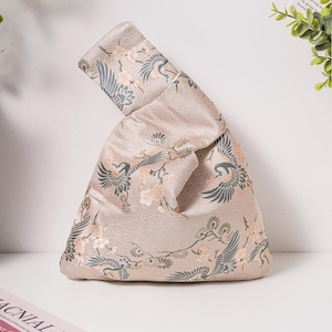 May include: A beige Japanese knot bag featuring a crane and floral design. The bag is made from a silky fabric and has a distinctive knotted structure. The cranes are grey, and the flowers are peach-coloured.