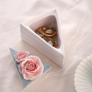 Könnte beinhalten: Eine Keramik-Schmuckschatulle in Dreiecksform mit einem gestreiften Design in Rosa und Blau. Die Schachtel ist geöffnet und enthält drei kleine goldfarbene gerahmte Stücke mit Blumendesigns. Im Vordergrund befindet sich eine kleinere Keramik-Schale in Dreiecksform mit einer blauen Glasur und zwei rosa Rosen.