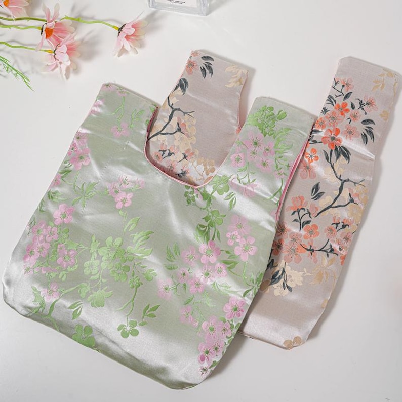 May include: Three satin tote bags with floral patterns. The bags feature a light green, pink, and peach colour scheme. The bags have a Japanese knot bag design, with a handle on each side. The floral patterns include pink and green blossoms.