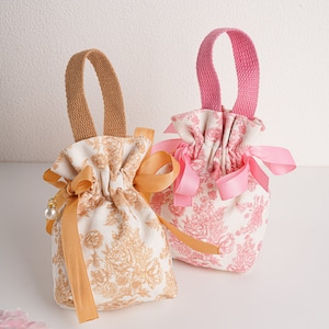Könnte beinhalten: Zwei kleine Stoffbeutel mit Kordelzug und Henkeln. Der Beutel links hat ein goldenes Blumenmuster und ein gelbes Band. Der Beutel rechts hat ein rosafarbenes Blumenmuster und ein rosafarbenes Band.