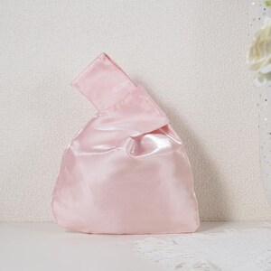 May include: A light pink satin knot bag with a smooth, reflective finish. The handbag features a single integrated handle formed by pulling one side of the fabric through the other, creating a soft, rounded silhouette with a subtle sheen.