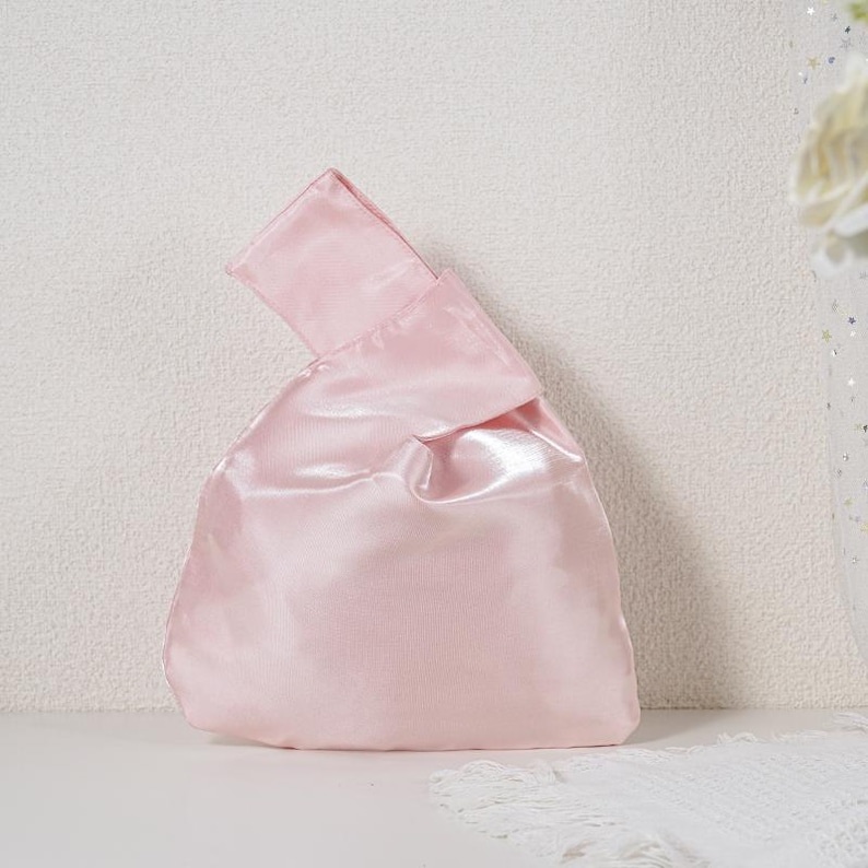 May include: A light pink satin knot bag with a smooth, reflective finish. The handbag features a single integrated handle formed by pulling one side of the fabric through the other, creating a soft, rounded silhouette with a subtle sheen.
