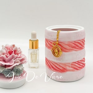 Op de afbeelding: Een witte keramische pot met roze en witte strepen, een gouden ketting met een gouden hanger, een klein doorzichtig glazen flesje met een gouden dop en een roze en witte bloem op een witte basis met de tekst "Joie De Vivre".