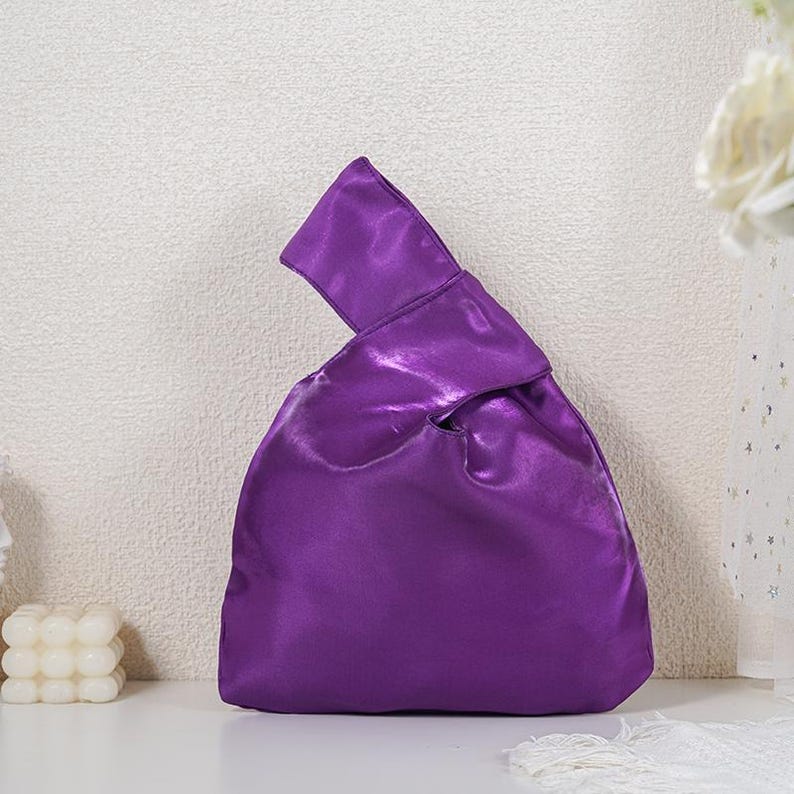 May include: A vibrant purple satin handbag featuring a distinctive Japanese knot design where one handle is longer than the other, allowing it to loop through for a secure closure. The fabric has a smooth, reflective sheen and a soft structure.