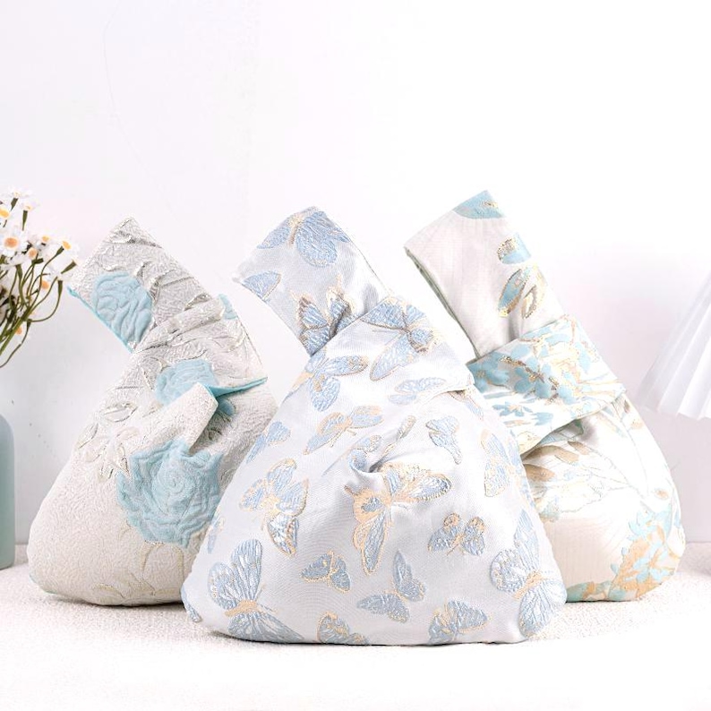 May include: Three decorative knot bags in a row. The bags are white with blue and gold butterfly patterns. The bag on the left has a floral design. The bags are made of a textured fabric.