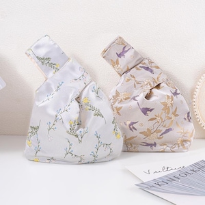 May include: Two fabric knot bags with floral and bird patterns. One bag is light grey with blue and yellow flowers. The other bag is beige with gold flowers and purple birds. The bags are on a white surface.