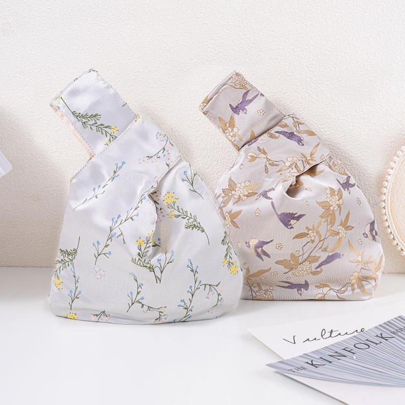 May include: Two fabric knot bags with floral and bird patterns. One bag is light grey with blue and yellow flowers. The other bag is beige with gold flowers and purple birds. The bags are on a white surface.