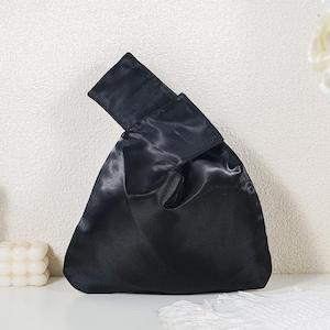 May include: A black satin handbag featuring a soft, unstructured silhouette with a single integrated handle loop. The fabric has a smooth, reflective sheen, and the design includes a folded top closure that creates a triangular shape when held.