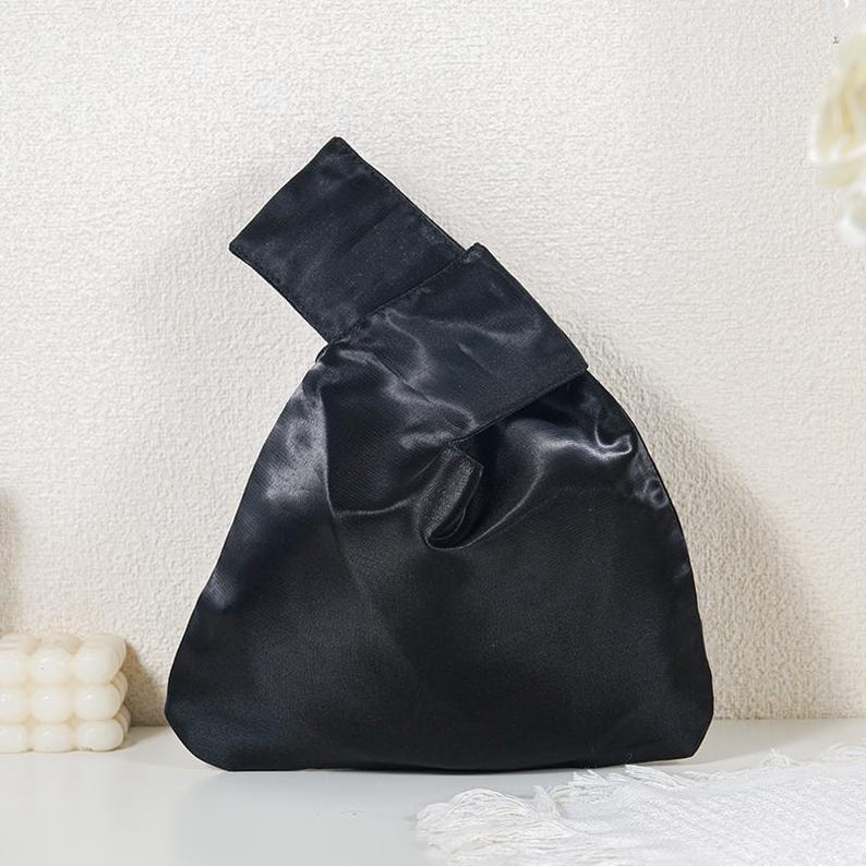 May include: A black satin handbag featuring a soft, unstructured silhouette with a single integrated handle loop. The fabric has a smooth, reflective sheen, and the design includes a folded top closure that creates a triangular shape when held.