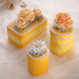 Op de afbeelding: Drie keramische containers met bloemmotieven. De containers zijn geel en wit met een gestreept patroon. De bloemen zijn van porselein en zijn in tinten geel en wit.