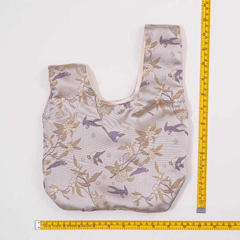 May include: A pale grey tote bag with a bird and floral design. The bag has a single handle and is approximately 30 cm wide. The pattern includes birds and flowering branches in brown, purple, and cream tones.