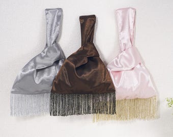May include: Three satin knot bags with a single handle and fringe trim at the bottom. The bags are colored silver with silver fringe, dark brown with black fringe, and light pink with gold fringe, each featuring a smooth, lustrous fabric finish.