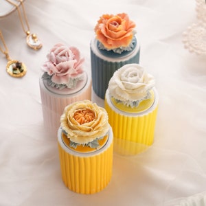 Op de afbeelding: Vier keramische potten met bloemendeksels. De potten zijn in tinten roze, geel en blauw. Elke pot heeft een andere bloem bovenop, waaronder een roze roos, een witte roos, een oranje roos en een gele roos.
