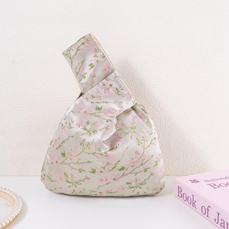 May include: A light grey knotted handbag featuring a floral pattern of pink blossoms and green leaves. The bag has a rectangular handle and a soft, rounded body. The bag is on a white surface.