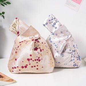 Può includere: Due borse a nodo realizzate in tessuto luccicante. La borsa a sinistra è rosa chiaro con fiori di ciliegio ricamati in rosa scuro e rami dorati. La borsa a destra è bianca con motivi di fiori di ciliegio blu e rami argentati.