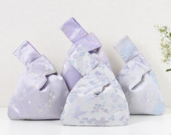 May include: Three Japanese-style knot bags made of satin fabric in shades of light purple and white. Each bag features a unique floral pattern with a subtle sheen and a single handle that loops through the other to close the pouch securely.