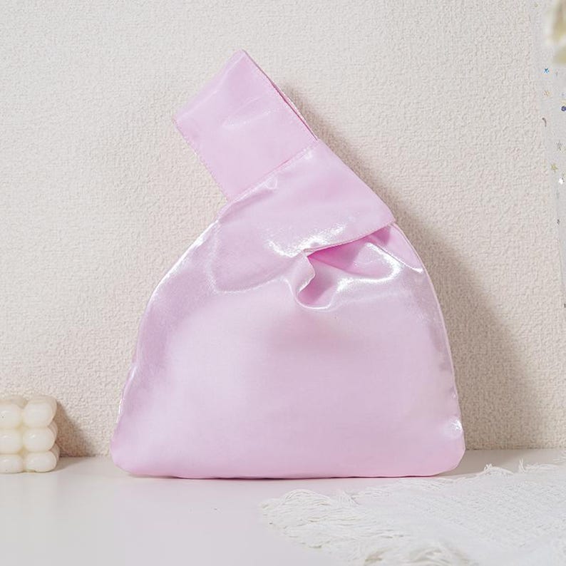 May include: A light pink satin handbag featuring a single integrated handle design. The bag has a smooth, reflective surface with a soft sheen and a triangular silhouette, created by pulling one handle through the other to close the opening.