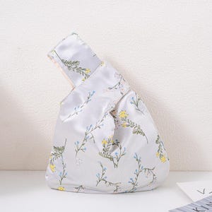 May include: A pale grey satin knot bag with a floral design. The bag has a repeating pattern of yellow, blue, and green flowers and leaves on slender stems. The handle is folded over and secured with a knot.