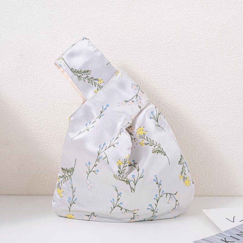 May include: A pale grey satin knot bag with a floral design. The bag has a repeating pattern of yellow, blue, and green flowers and leaves on slender stems. The handle is folded over and secured with a knot.